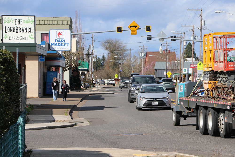 A busy street with businesses, pedestrians walking on the sidewalk, and midday vehicle traffic. 