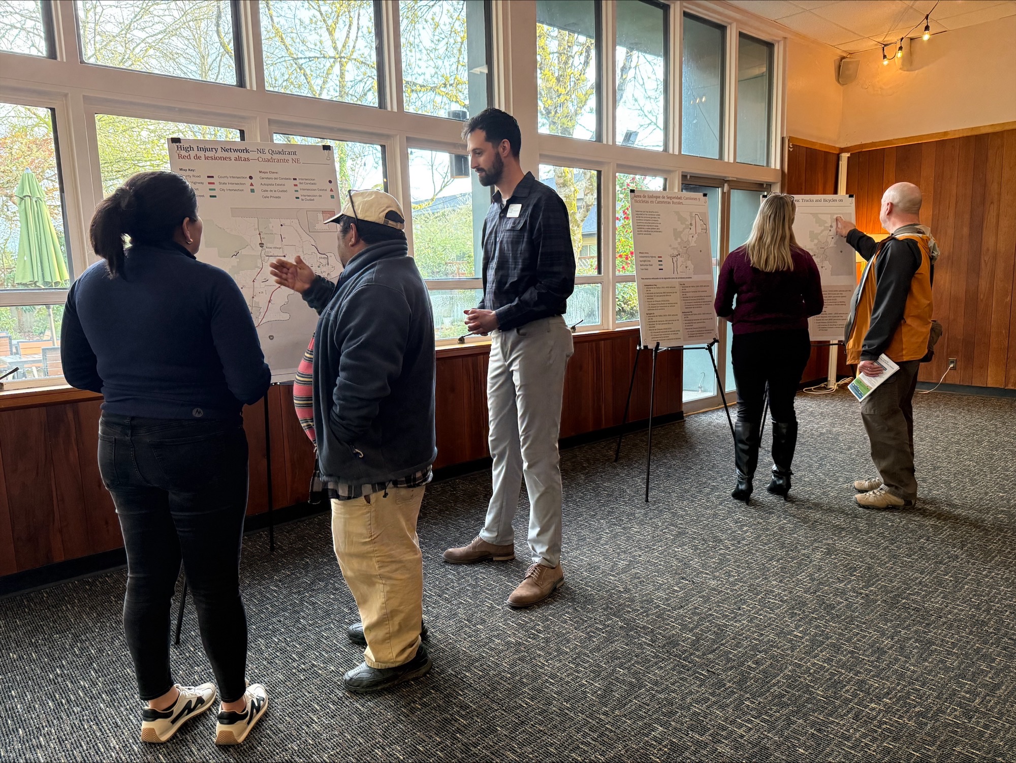 County staff and project staff members talk with members of the public during an open house event. Display boards with maps of the project high injury network locations are shown.