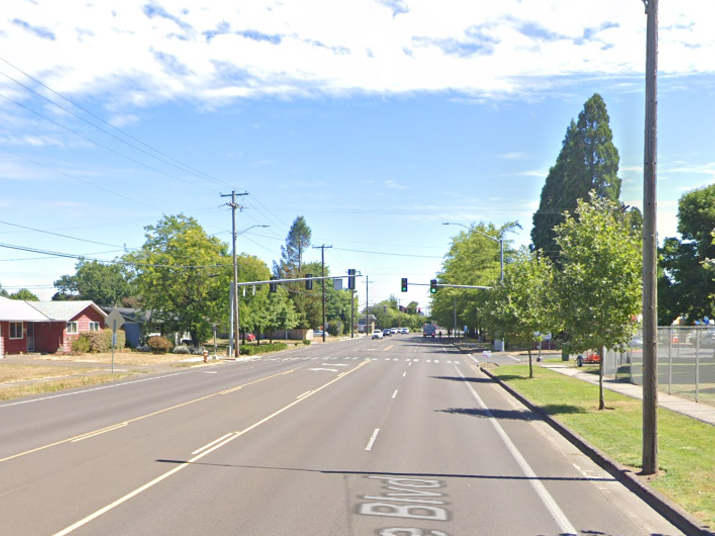 A four-lane street leading to an intersection with overhead traffic lights on a sunny day.  