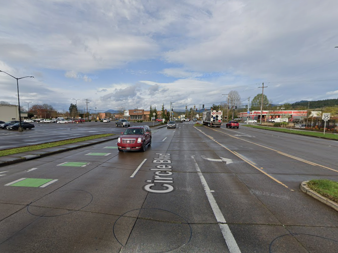 A wide six-lane street on an overcast day, with a red SUV driving next to a green-striped bike lane.  