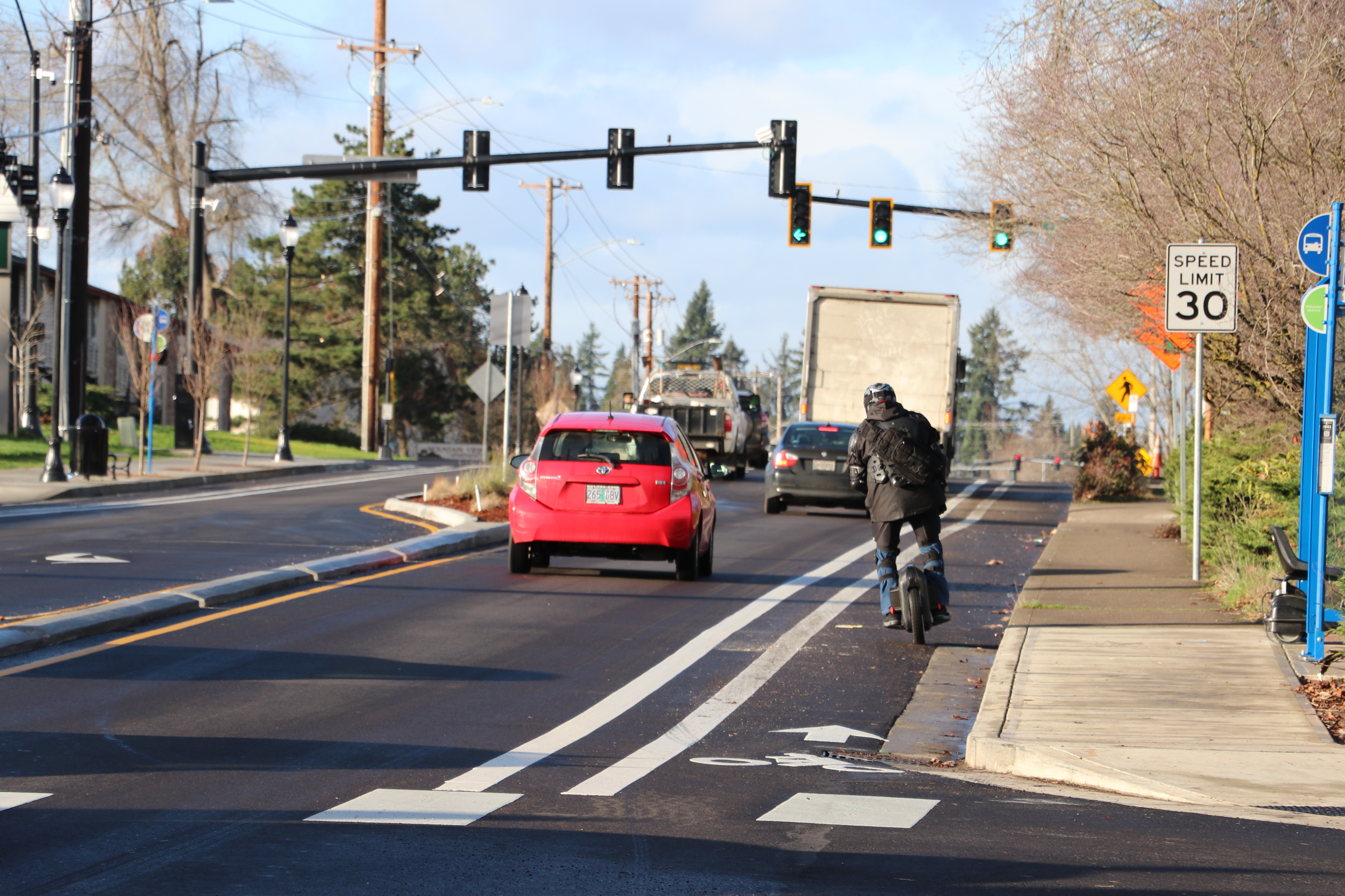 Cars traveling toward a stoplight, and a cyclist is traveling in a bike lane. A sign reads: Speed Limit 30. 